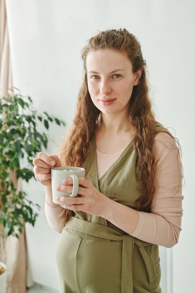 Pregnant woman smiling while holding a mug, standing in a bright indoor setting.