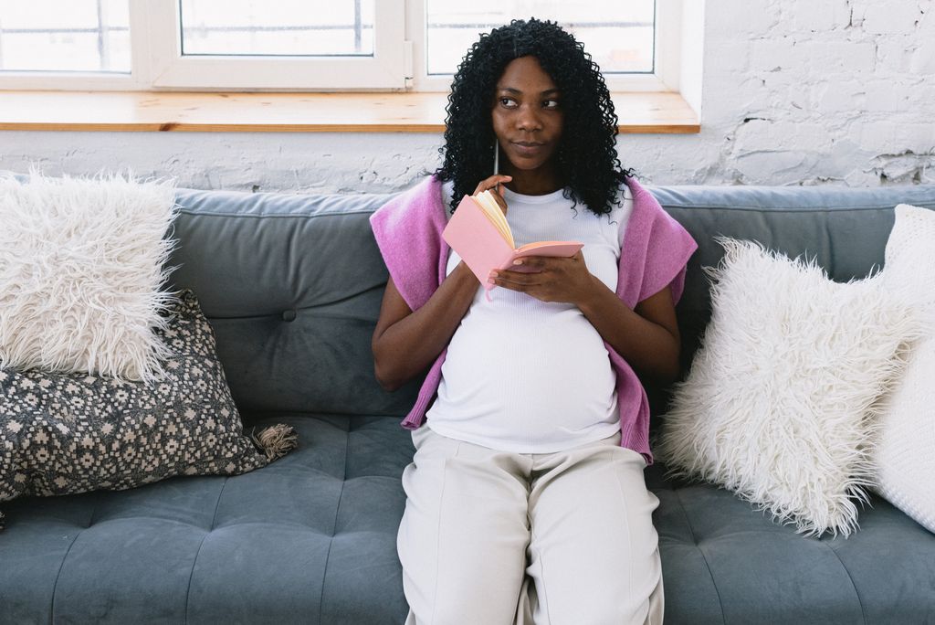 Thoughtful pregnant African American female taking notes in notepad and looking away while sitting on comfortable couch in living room