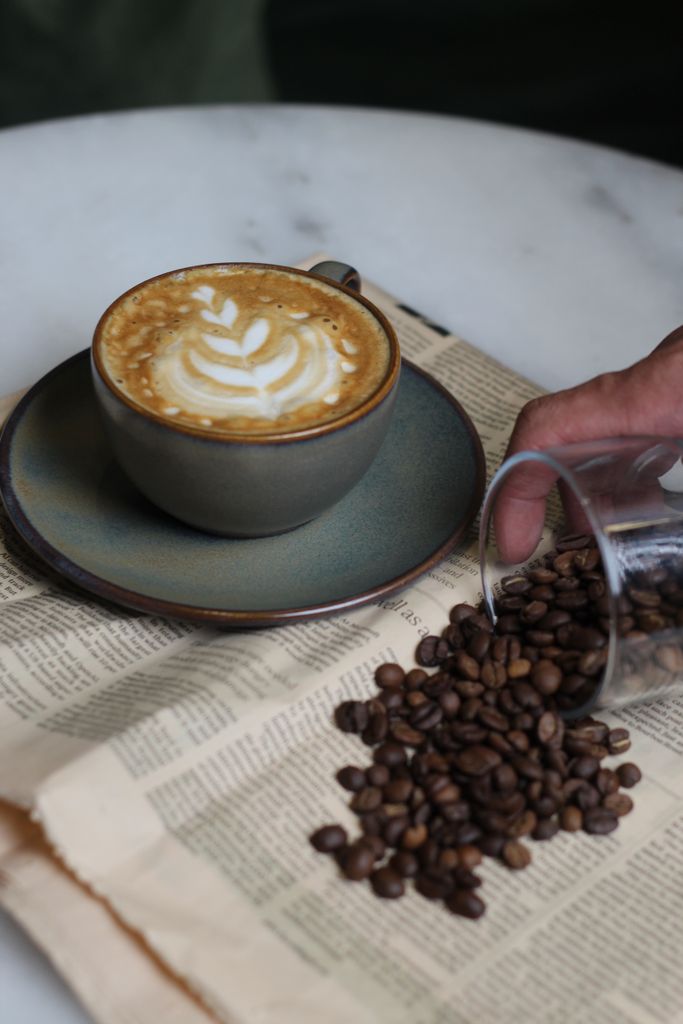 A cappuccino with latte art next to spilled coffee beans on a vintage newspaper.