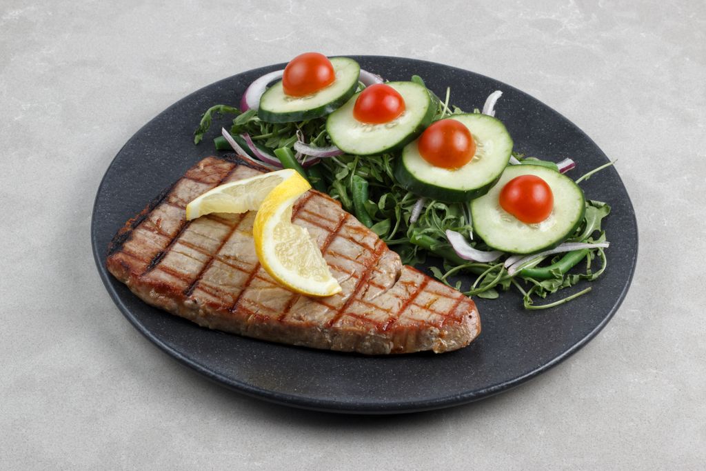 A grilled tuna steak topped with lemon slices served with a fresh cucumber and tomato salad on a dark plate.