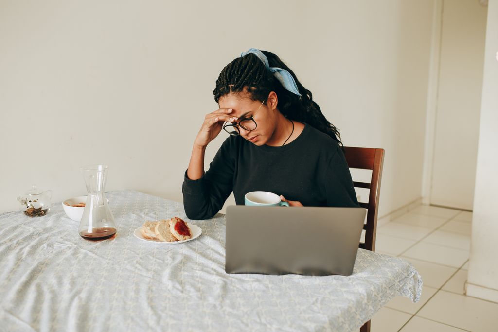 Woman working from home, stressed, drinking coffee, seated at table with laptop.